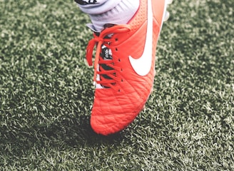 A close-up view of a red soccer cleat with white laces and a white logo on green artificial turf. The shoe is worn by a person whose sock is visible above the shoe.