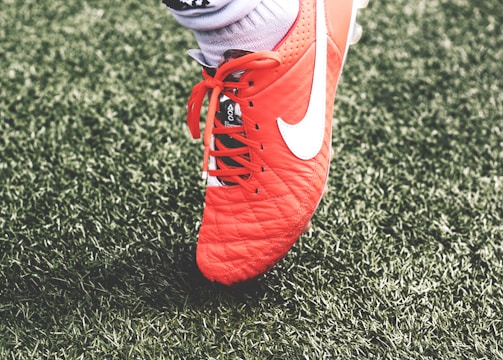 A close-up view of a red soccer cleat with white laces and a white logo on green artificial turf. The shoe is worn by a person whose sock is visible above the shoe.