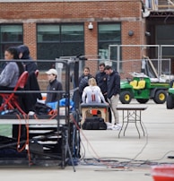 A group of people are gathered around an individual wearing a sports jersey with the number 11. They appear to be in a sports facility area, possibly a training or medical setup. There are tables, chairs, and equipment scattered around, with a brick building in the background. Some individuals are seated on a platform with electronics close by, suggesting a recording or monitoring activity. Utility vehicles and an orange barrier are present in the background.