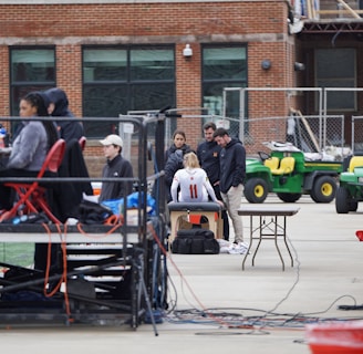 A group of people are gathered around an individual wearing a sports jersey with the number 11. They appear to be in a sports facility area, possibly a training or medical setup. There are tables, chairs, and equipment scattered around, with a brick building in the background. Some individuals are seated on a platform with electronics close by, suggesting a recording or monitoring activity. Utility vehicles and an orange barrier are present in the background.