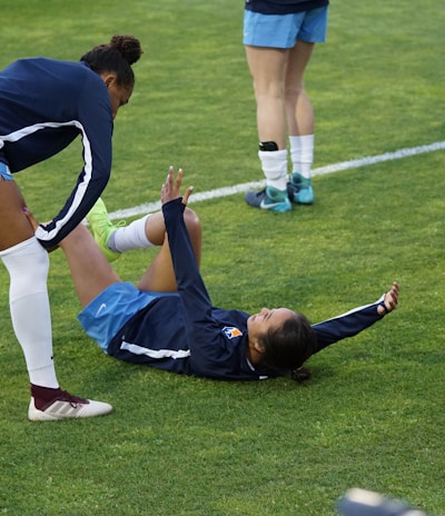 A group of athletes in sportswear are on a green soccer field. Two individuals are interacting closely; one helps the other who is lying on the grass. Nearby, other athletes are stretching.
