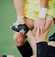 A close-up of a sports interaction where one person in a yellow uniform helps another wearing black leg gear. The person in yellow is gripping the leg of the other individual, providing support. The background appears to be a green sports field.