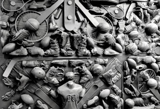 A complex arrangement of various sports equipment and attire, including tennis rackets, soccer balls, basketballs, helmets, shoes, gloves, and a mannequin torso wearing a T-shirt. The items are layered and artistically displayed in a monochrome color scheme, creating an intricate texture and visual pattern.