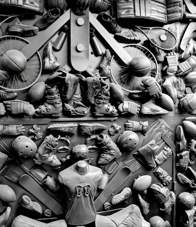 A complex arrangement of various sports equipment and attire, including tennis rackets, soccer balls, basketballs, helmets, shoes, gloves, and a mannequin torso wearing a T-shirt. The items are layered and artistically displayed in a monochrome color scheme, creating an intricate texture and visual pattern.