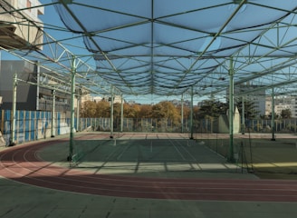 A spacious indoor sports facility with a tennis court and running tracks. The court is surrounded by high nets and tall, green metal structures supporting a roof. The area is well-lit with natural light filtering in through a transparent ceiling. In the background, there are trees with autumn foliage and several residential buildings.