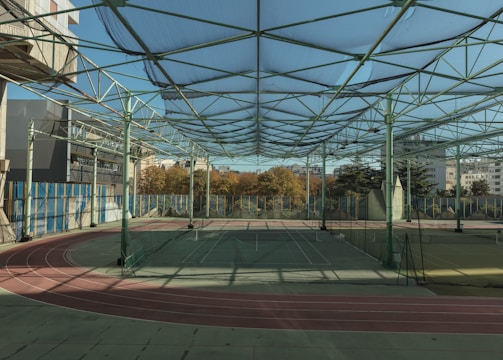 A spacious indoor sports facility with a tennis court and running tracks. The court is surrounded by high nets and tall, green metal structures supporting a roof. The area is well-lit with natural light filtering in through a transparent ceiling. In the background, there are trees with autumn foliage and several residential buildings.