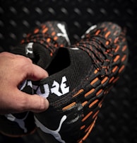 A hand holding a pair of black sports shoes featuring a white logo and the word 'FUTURE' in bold letters. The shoes have orange accents and textured patterns on a dark, patterned background.