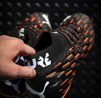 A hand holding a pair of black sports shoes featuring a white logo and the word 'FUTURE' in bold letters. The shoes have orange accents and textured patterns on a dark, patterned background.