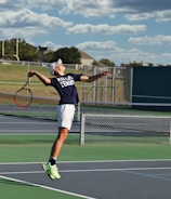 A tennis player is captured mid-air while serving at an outdoor tennis court. The player is wearing a navy blue shirt with 'Keller Tennis' printed on it, white shorts, and bright green sports shoes. A tennis racket is in the player's right hand, while the tennis ball is in the air above. The background includes a chain-link fence, a field, trees, and a partly cloudy sky.