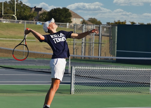 A tennis player is captured mid-air while serving at an outdoor tennis court. The player is wearing a navy blue shirt with 'Keller Tennis' printed on it, white shorts, and bright green sports shoes. A tennis racket is in the player's right hand, while the tennis ball is in the air above. The background includes a chain-link fence, a field, trees, and a partly cloudy sky.