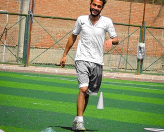 A person in sports clothing is running on a green artificial turf with white and red cones arranged in a pattern. The individual appears to be participating in a sports drill or exercise. The background includes a brick wall and netting consistent with an outdoor sports facility.