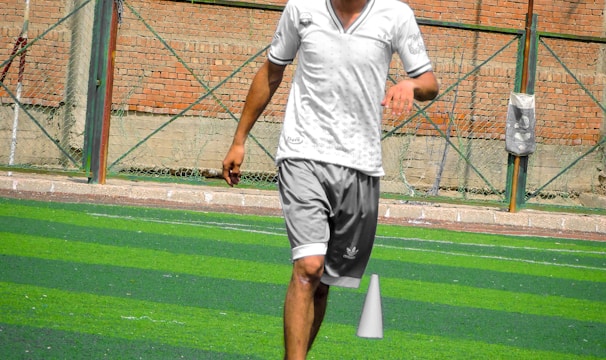 A person in sports clothing is running on a green artificial turf with white and red cones arranged in a pattern. The individual appears to be participating in a sports drill or exercise. The background includes a brick wall and netting consistent with an outdoor sports facility.