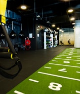 A modern gym space featuring a green track laid out on the floor with numbered lanes. TRX suspension trainers hang prominently in the foreground. The gym is equipped with various exercise equipment, including kettlebells and punching bags. In the background, there is a FIT sign near a reception desk under a low-ceilinged industrial design.