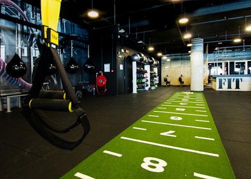 A modern gym space featuring a green track laid out on the floor with numbered lanes. TRX suspension trainers hang prominently in the foreground. The gym is equipped with various exercise equipment, including kettlebells and punching bags. In the background, there is a FIT sign near a reception desk under a low-ceilinged industrial design.