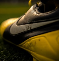 A close-up view of a football or soccer shoe with a distinctive swoosh logo in focus, featuring scuffed and worn textures on the leather surface. The shoe is predominantly black and yellow, with rich detailing captured under gleaming light. The background includes blurred green artificial turf, suggesting a sports field setting.