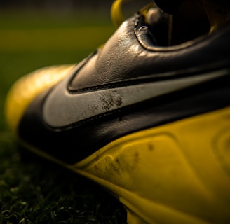 A close-up view of a football or soccer shoe with a distinctive swoosh logo in focus, featuring scuffed and worn textures on the leather surface. The shoe is predominantly black and yellow, with rich detailing captured under gleaming light. The background includes blurred green artificial turf, suggesting a sports field setting.