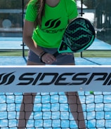 A person stands on a padel court, holding a black padel racket with their body slightly bent forward. They are wearing a bright green shirt with the word 'Sidespin' printed on it, along with gray shorts. The blue court surface is visible, and there is a net in the foreground with the 'Sidespin' logo.