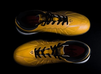A pair of yellow sports shoes with black laces is placed against a dark background. The design is sleek, with angular patterns on the surface. The inside of the shoes appears to have a red lining with a brand logo visible.