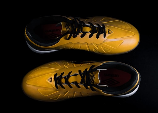A pair of yellow sports shoes with black laces is placed against a dark background. The design is sleek, with angular patterns on the surface. The inside of the shoes appears to have a red lining with a brand logo visible.