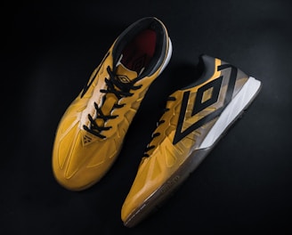 A pair of yellow and black sports shoes displayed against a dark background. The shoes feature a bold geometric pattern and are designed for athletic performance.