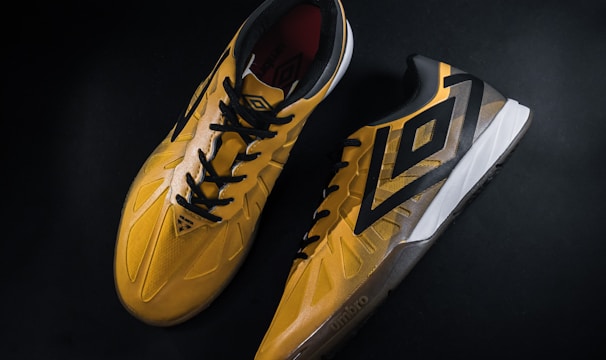A pair of yellow and black sports shoes displayed against a dark background. The shoes feature a bold geometric pattern and are designed for athletic performance.