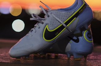 A pair of white soccer cleats with yellow and black detailing is propped on a surface against a backdrop of a vibrant sunset. The cleats are well-lit, showcasing their design and brand logo prominently.