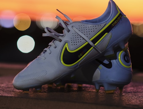 A pair of white soccer cleats with yellow and black detailing is propped on a surface against a backdrop of a vibrant sunset. The cleats are well-lit, showcasing their design and brand logo prominently.