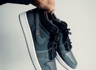 A hand is holding a pair of dark-colored athletic shoes. The shoes have a combination of leather and mesh materials with a swoosh logo on the side. They feature black laces and white soles. The hand appears to be wearing a black sleeve.