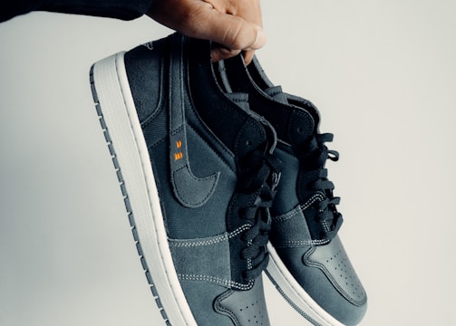 A hand is holding a pair of dark-colored athletic shoes. The shoes have a combination of leather and mesh materials with a swoosh logo on the side. They feature black laces and white soles. The hand appears to be wearing a black sleeve.