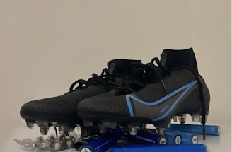 A pair of black and blue soccer cleats with metal studs, placed on top of a blister pack of capsules and several blue vape pens. The cleats have a sleek design with blue accents and appear to be of professional quality.