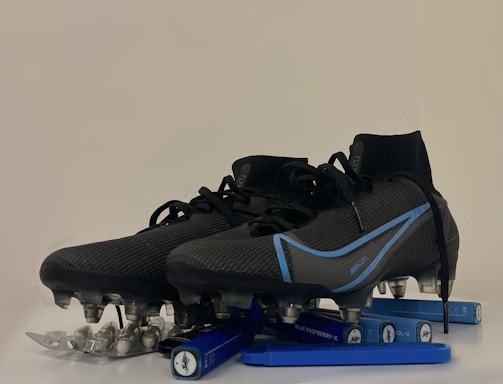 A pair of black and blue soccer cleats with metal studs, placed on top of a blister pack of capsules and several blue vape pens. The cleats have a sleek design with blue accents and appear to be of professional quality.