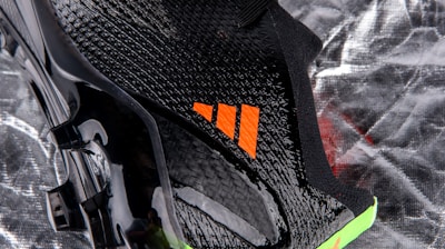 A close-up view of a sleek black sports shoe with a textured surface and an orange logo. The sole has a shiny finish with molded studs, and there is a neon green accent at the bottom.