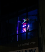 A neon sign in a window displays vibrant colors with tennis sporting motifs and text, which appears above a business sign advertising sporting goods and music. The setting is dimly lit, enhancing the glow of the neon signage.