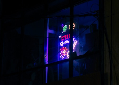 A neon sign in a window displays vibrant colors with tennis sporting motifs and text, which appears above a business sign advertising sporting goods and music. The setting is dimly lit, enhancing the glow of the neon signage.